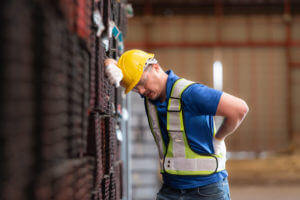 A male worker wearing a safety vest and helmet is standing on a steel pallet due to back and spine pain from lifting heavy objects in a factory.