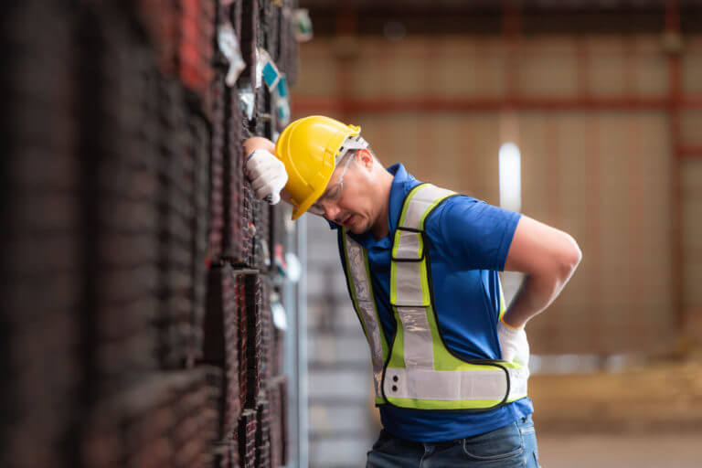 A male worker wearing a safety vest and helmet is standing on a steel pallet due to back and spine pain from lifting heavy objects in a factory.
