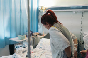 A young woman seen from behind adjusting her hospital gown in a hospital room for back surgery.