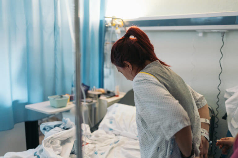 A young woman seen from behind adjusting her hospital gown in a hospital room for back surgery.