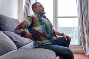 Mature man suffering from back pain while sitting on sofa at home due to his herniated disc.